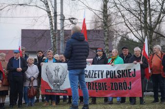 Przemawiający mężczyzna, w tle grupa osób z polskimi flagami oraz transparentem na którym widnieje polski orzeł oraz napis Strefa wolnego słowa gazety polskiej Jastrzębie-Zdrój