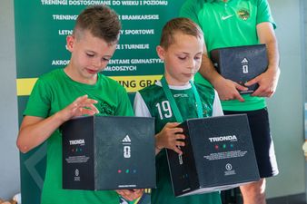 Dwóch młodych chłopców w zielonych koszulkach piłkarskich z uwagą ogląda czarne pudełka z logo Adidas i napisem „TRIONDA”, które trzymają w rękach