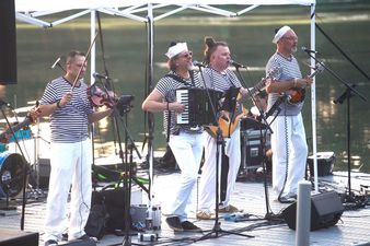 Zespół szantowy grający koncert na pomoście przy brzegu jeziora. Muzycy w strojach marynarskich wykonują utwór na instrumentach strunowych i akordeonie