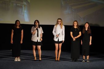 trzy dziewczyny w czarnych strojach między nimi dwie w czarnych spoódnicach i białych bluzkach koszulowych śpiewają