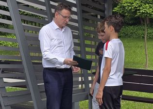 Prezydent wręcza dyplom dwóm chłopcom, którzy mu się przyglądają. Wszyscy stoją pod nowoczesnym zadaszeniem.