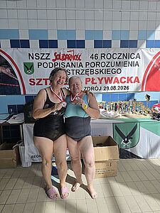 Dwie uśmiechnięte kobiety w strojach kąpielowych i z medalami na szyi obejmują się na tle baneru okolicznościowego i trofeów
