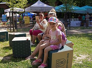 Mala dziewczynka i dwie dorosłe siedzą na kostkach z herbem i napisem Jastrzębie-Zdrój, w tle kolorowe namioty