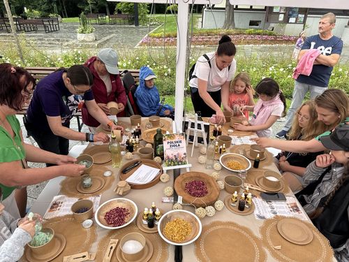 Uczestnicy pikniku, dorośli i dzieci, zgromadzeni wokół stołu warsztatowego z suszonymi ziołami, olejkami i miskami z płatkami kwiatów. W tle widoczna tabliczka z napisem „Polskie zioła”
