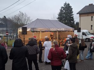 Widok na świąteczne wydarzenie plenerowe Sołectwa Bzie. Publiczność i osoby w strojach aniołów stoją przed dużym imprezowym namiotem. W tle budynek, biały bus i wysoki świerk