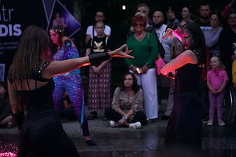 dwoje artystów w ciemnych strojach ciemnych charakterów wykonuje ekspresyjne gesty dłońmi podczas wieczornego spektaklu plenerowego w czerwonym świetle