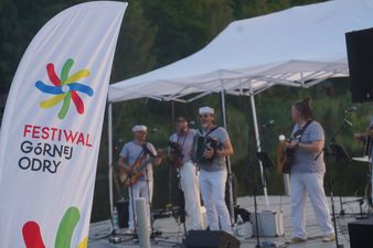 Wielobarwna flaga z logo i napisem Festiwal Górnej Odry na pierwszym planie. W tle, pod białym namiotem, występuje zespół muzyczny w marynarskich strojach