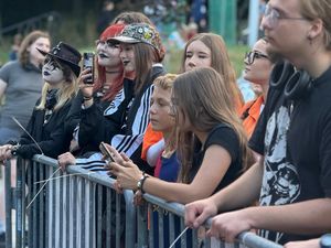 12. Grupa ludzi stoi za barierkami pod sceną patrząc co się na niej dzieje. W tle widać przechodzące osoby. 