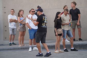 Mężczyzna w czapce z daszkiem żywo gestykulujący podczas rozmowy w tłumie uczestników imprezy plenerowej
