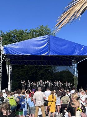 Na scenie z niebieskim zadaszeniem występuje grupa artystów w ciemnych strojach, a przed sceną zgromadziła się liczna publiczność