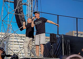 Dwóch mężczyzn śpiewa do mikrofonu na scenie, jeden z nich ma uniesione ramię