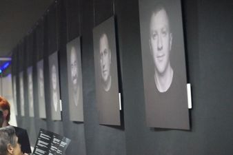 Na czarnej ścianie wisi kolekcja zdjęć portretowych mężczyzn, z boku kilka kobiet oglądających zdjęcia