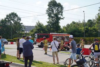 Grupa dorosłych i rowerzystów obserwuje akcję strażaków przy uszkodzonym aucie. W tle widoczny jest wóz strażacki i drzewa
