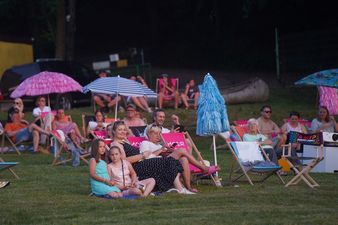 Grupa widzów siedząca na leżakach i ręcznikach na trawie, niektórzy pod parasolami