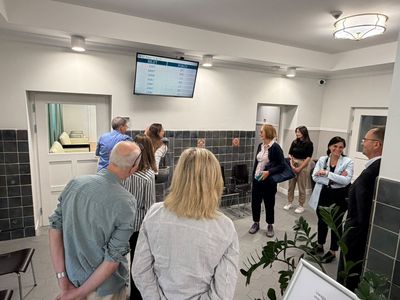 Grupa osób w poczekalni urzędu; na ścianie z ciemnych kafelków wisi ekran systemu kolejkowego
