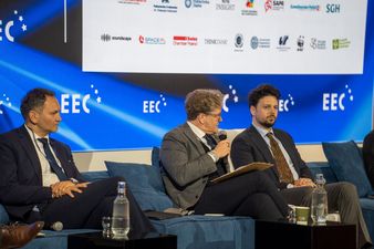 Trzech mężczyzn w garniturach na scenie w tle ekran z napisiem EEC i trzema białymi gwiazdami