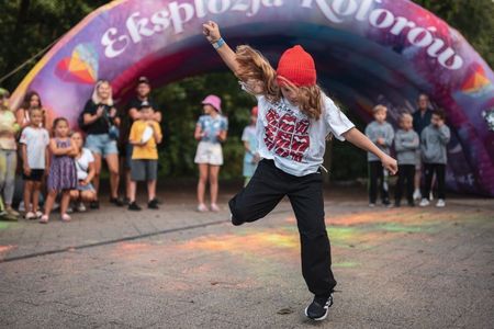 Dziewczynka w czerwonej czapce tańczy breakdance na kostce brukowej przed dużą, dmuchaną bramą festiwalową