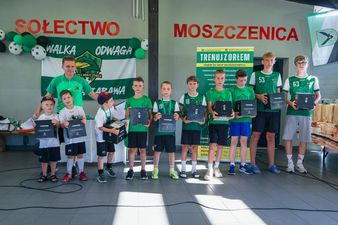 Trenerzy oraz grupa młodych piłkarzy w różnych kategoriach wiekowych pozują do zdjęcia, trzymając w rękach otrzymane nagrody w czarnych pudełkach, z banerem reklamowym klubu „Orzeł Moszczenica” w tle