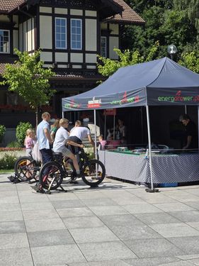 Uczestnicy festynu pedałują na rowerach stacjonarnych pod namiotem „energorower.pl” ustawionym obok stylowego budynku