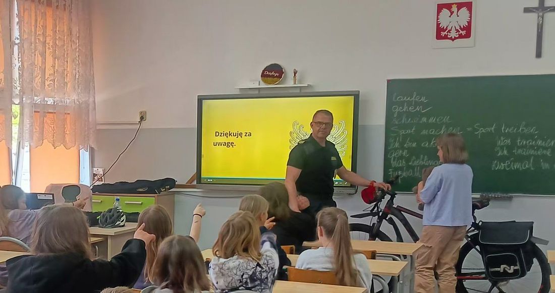 Strażnik podczas prelekcji trzymający rower, za nim ekran z prezentacją. Obok niego zapisana kredą tablica