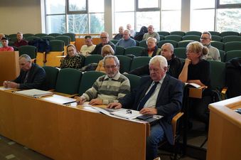 Grupa kilkunastu osób siedzi w ławkach sali obrad i słucha prezentacji