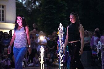 dwie performerki w błyszczących strojach spacerują wieczorem między podświetlonymi strumieniami miejskiej fontanny posadzkowej przed widownią