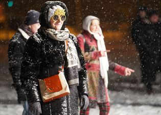 Starsza kobieta ubrana w pozłacane okulary przeciwsłoneczne z napisem Happy New Year. Wokół padający śnieg.