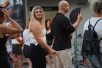 Blondynka w białym topie i mężczyzna z wachlarzem wśród uczestników plenerowego wydarzenia