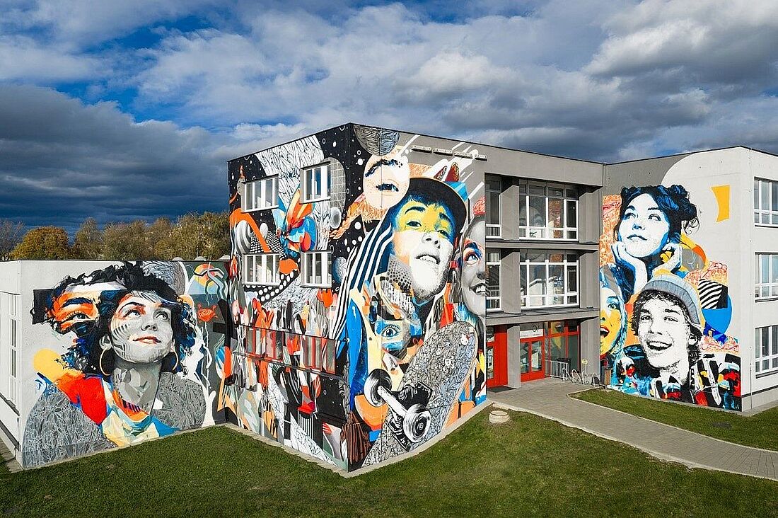 Mural pt. „Pęd życia” na ścianach budynku przedstawia dynamiczną, kolorową kompozycję z twarzami młodych ludzi, deskorolką i wzorami graficznymi