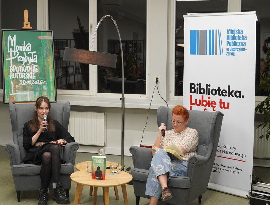 dwa szare fotele na których siedzą dwie kobiety jedna rudowłosa, druga burnetka na stoliku między fotelami ksiażka autorki w tle rollup Biblioteki oraz afisz spotkania autorskiego