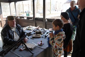 Dzieci i dorośli słuchający wyjaśnień mężczyzny w mundurze, przed nimi na stole zabytkowa broń