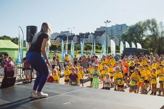 Kobieta na scenie prezentuje ćwiczenia zebranym dzieciom, w tle bloki i domki mieszkalne