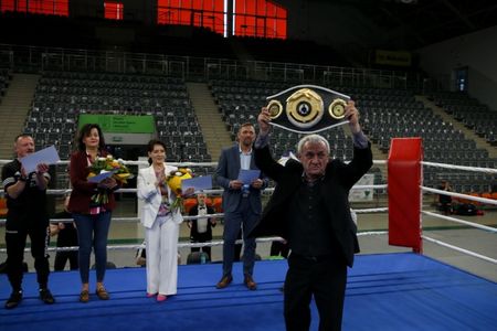mężczyzna w czarnym garniturze unosi bokserski pas mistrzowski, stojąc w ringu. W tle na niebieskiej macie stoją oficjele z kwiatami na tle pustych trybun hali widowiskowo-sportowej