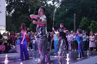 Grupa aktorów w połyskujących kostiumach występuje w wodzie rozświetlonej przez podłogowe reflektory fontanny na tle zgromadzonych widzów