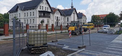 Prace brukarskie przed białym budynkiem z wieżyczką. Na pierwszym planie zbiornik DPPL, dalej koparka i żółty furgon. Teren robót oddzielony jest metalowym ogrodzeniem i słupkami