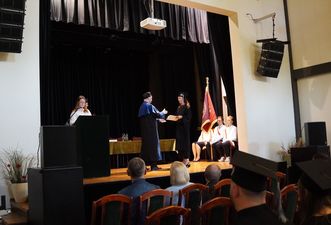 Uroczyste wręczenie karty absolutoryjnej studentce przez rektora na scenie. W tle widoczny poczet sztandarowy z flagą