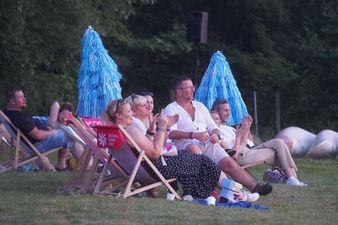 Publiczność festiwalowa siedzi wieczorem na leżakach i klaszcze w stronę sceny. Między rzędami stoją dekoracyjne, niebieskie parasole z frędzlami