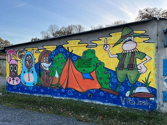 Kolorowy mural przedstawiający czechosłowackie postacie bajkowe: Żwirka i Muchomorka, Rumcajsa oraz lisa przy pomarańczowym namiocie