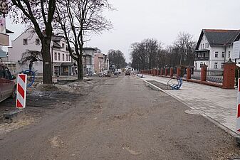 Widok wzdłuż szerokiej, gruntowej drogi w trakcie przebudowy, przy której ustawiono biało-czerwone słupki ostrzegawcze. Po prawej stronie widać gotowy fragment nowego chodnika oraz wystające z ziemi niebieskie rury instalacyjne, a po lewej stronie ulicy stoją starsze budynki i bezlistne drzewa