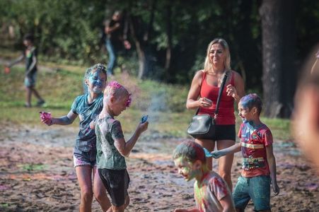 Dzieci umazane kolorowymi proszkami bawią się na piaszczystym terenie pod okiem stojącej w tle kobiety