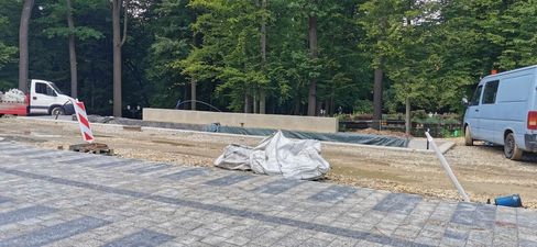 Plac budowy w parku. Na pierwszym planie ułożona kostka, dalej betonowe murki i zaparkowane furgonetki na tle ściany lasu