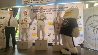 Trzy młode judoczki z medalami i dyplomami na podium podczas turnieju w Prievidzy, obok sędzia oraz klubowa maskotka orła