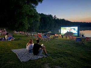 Seans kina plenerowego o zmierzchu – ludzie na leżakach i kocach oglądają film na dużym ekranie nad jeziorem
