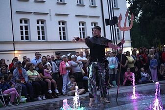 Aktor w ciemnym pancerzu i srebrnych spodniach stoi w fontannie, trzymając w ręku duży trójząb i wykonując gest dłonią w stronę publiczności.
