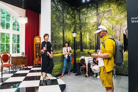 Grupa turystów zwiedza wystawę muzealną stylizowaną na dawny salon kurortowy. Po lewej stronie widać stylowe meble: zabytkowy zegar stojący, fotel oraz komodę na tle czerwonej kotary. Po prawej stronie znajduje się fototapeta przedstawiająca zielony park z latarnią, przy której stoją i odpoczywają uczestnicy spaceru