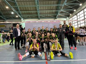 Brązowe młodziczki pozujące do zdjecia na tle siatki z trenerami i działaczami z okrzykiem radości