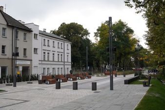 Nowo wybrukowany plac miejski z rzędem drewnianych ławek i betonowych słupków blokujących. W tle widoczne są kilkupiętrowe białe budynki oraz ściana zieleni