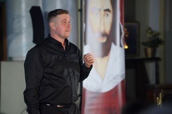 Z przodu mężczyzna ubrany na czarno utożsamiający księdza Jana Machy, w tle obraz z twarzą Jezusa Chrystusa