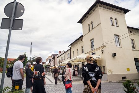 Uczestnicy spaceru z przewodnikiem idą deptakiem obok kawiarni z parasolami marki Kozel. W tle wznosi się jasny, zabytkowy budynek z charakterystyczną wieżą