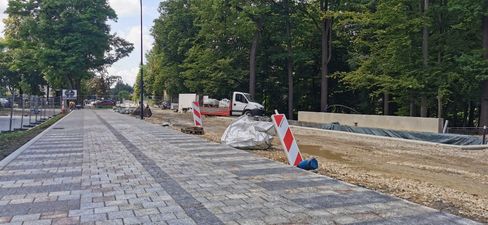 Ułożona alejka z kostki brukowej prowadząca w głąb parku. Obok trwają prace ziemne, widać białą ciężarówkę i pachołki na tle lasu